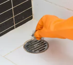 Tay mặc găng tay cao su màu cam đang lấy tóc ra khỏi nắp đậy cống thoát nước.